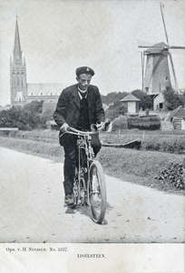 12334 Afbeelding van een jongeman op een rijwiel op de Hoge Biezen te IJsselstein, met op de achtergrond de R.K. St. ...